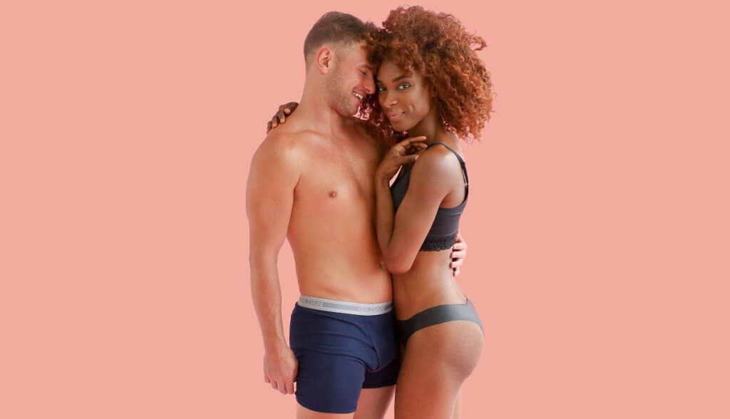 A white man wearing only boxers stands embracing a woman of color wearing a black bra and black Lorals panties.