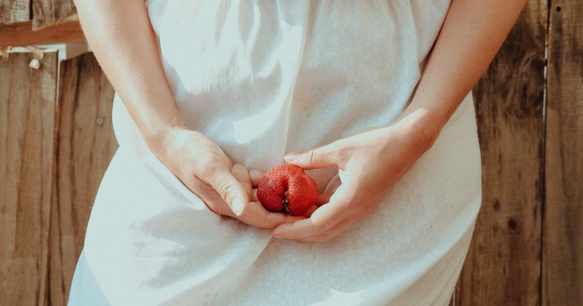 A woman wearing a white dress is shown holding a red fruit in front of her pelvic area.