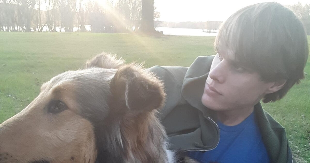 A young Caucasion man appeas sitting in a park next to a dog of the Collie breed.