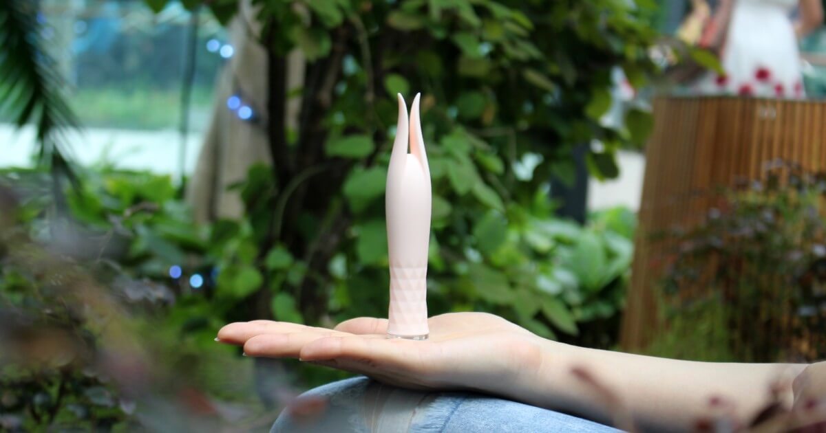 A light pink vibrator is held in the palm of person's hand.
