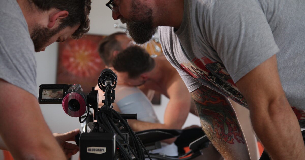 Two white men with beards are seen filming a male couple kissing, one of whom is quadripelgic and in a wheelchair.