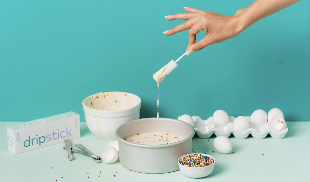 A hand holds a dripstick awkward essentials sex sponge that has batter on it, next to various baking equipment and foods.