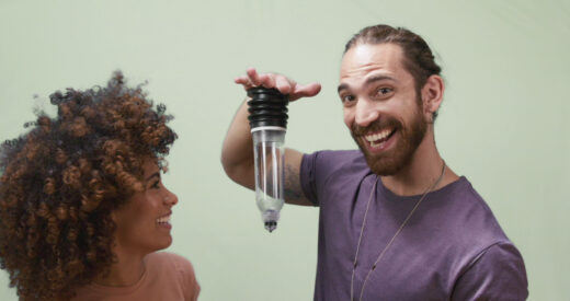 Man holds hand palm down, with an upside down Bathmate penis pump suctioned to his hand. A woman smiles looking at him.