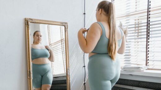 A young curvy woman stands in front of a mirror looking at herselg while wearing body-hugging undergarments.