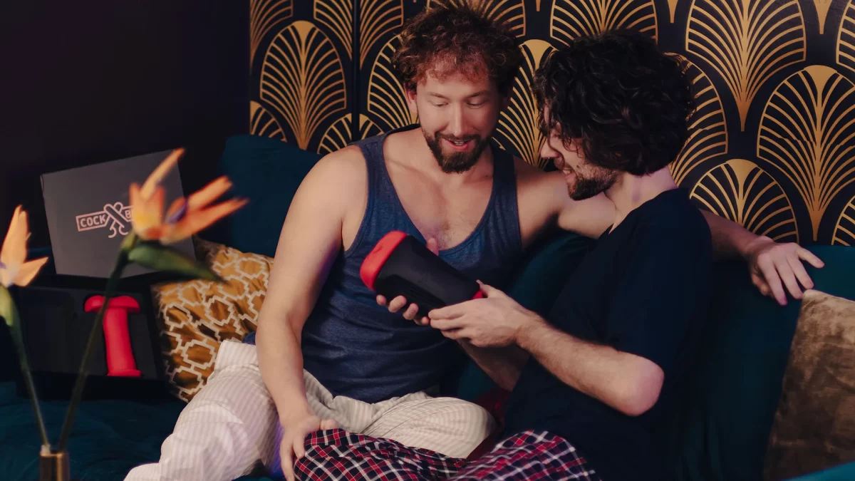 The gay frotting sex device Cock Block is held by a young gay man on a couch next to his male lover.